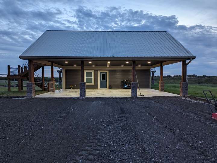 A picture of a house with a metal roof, with 4 wooden beams supporting it, there is then under the roof, a door, and a window to the left, and a window to the right,  and a concrete slab below it. Infront of the house, there is gravel that has not been compacted/finished. There is a semi blue greyish sky, that is a bit cloudy.