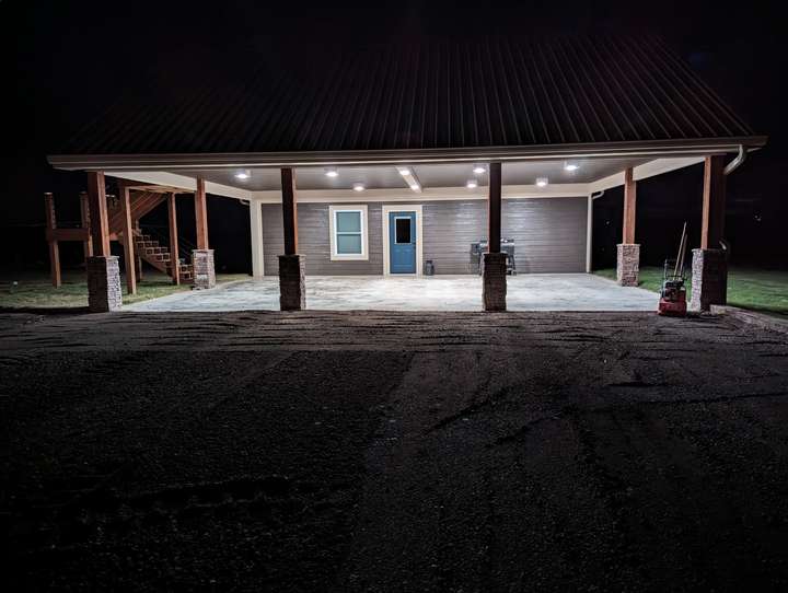 A picture of a house with a metal roof, with 4 wooden beams supporting it, there is then under the roof, a door, and a window to the left, and a window to the right,  and a concrete slab below it. Infront of the house, there is gravel that has not been compacted/finished. The sky is dark. It is at night.