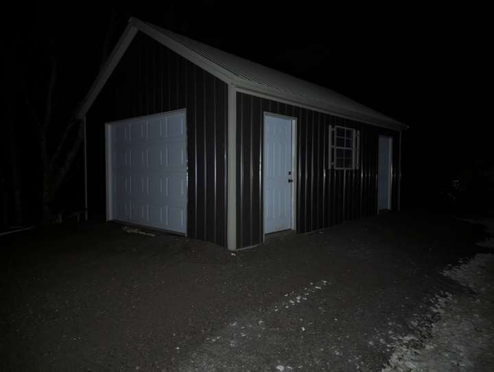 A picture of a grey shed constructed with metal with bends in it. There is a garage door in the front, and to the right there is a wall with two doors, and a window. there is grey material there. 